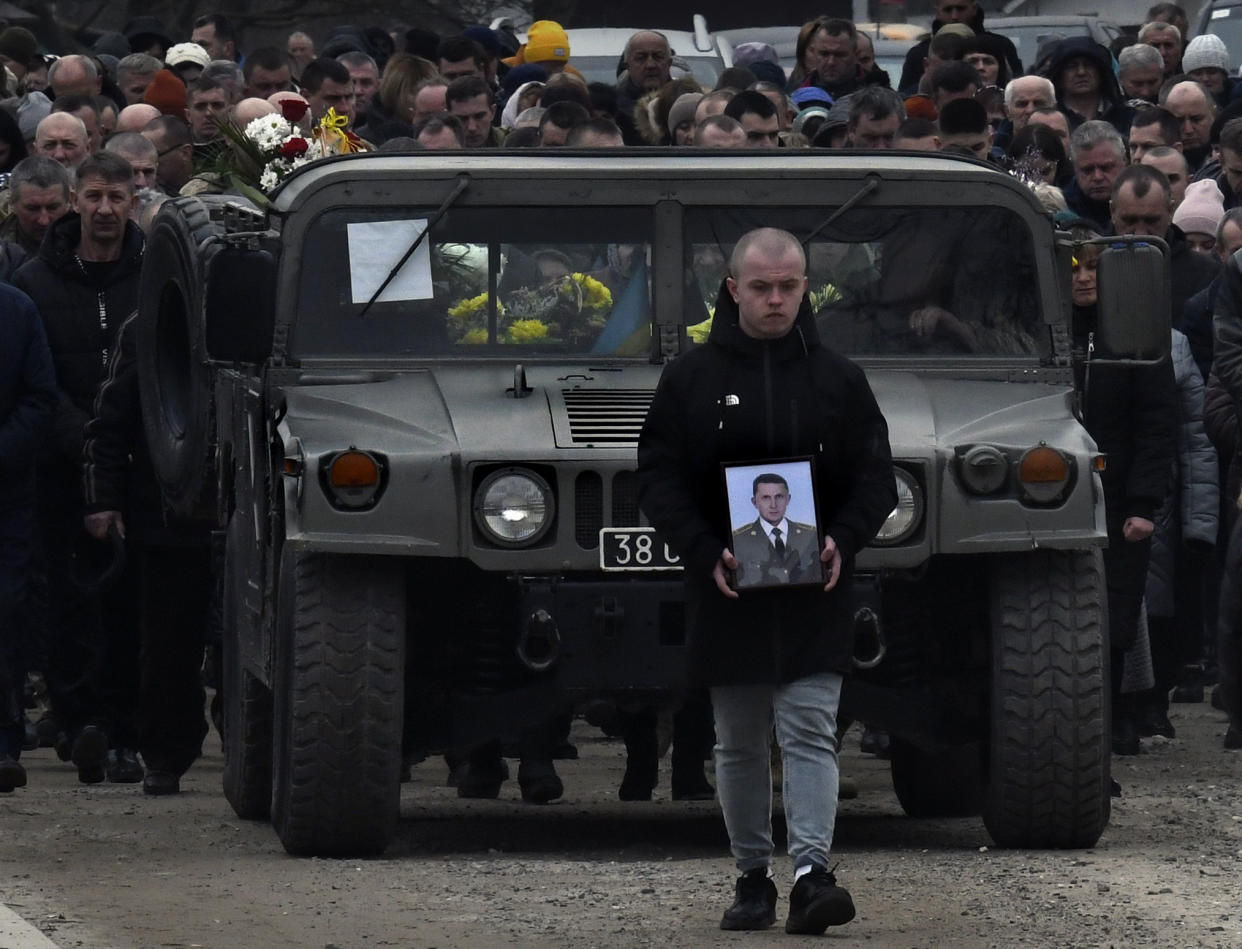 A man holding the picture of one of the fallen soldiers is followed by a procession of mourners.