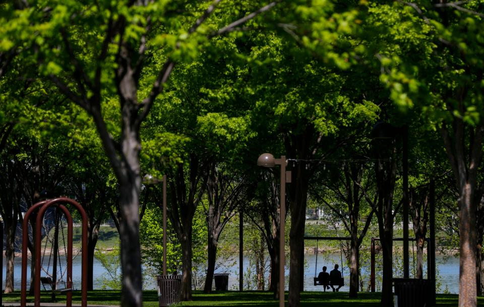 Two people sit on a swing under the trees at Waterfront Park along the Ohio River in Louisville on a sunny spring Wednesday afternoon. April 19, 2023
