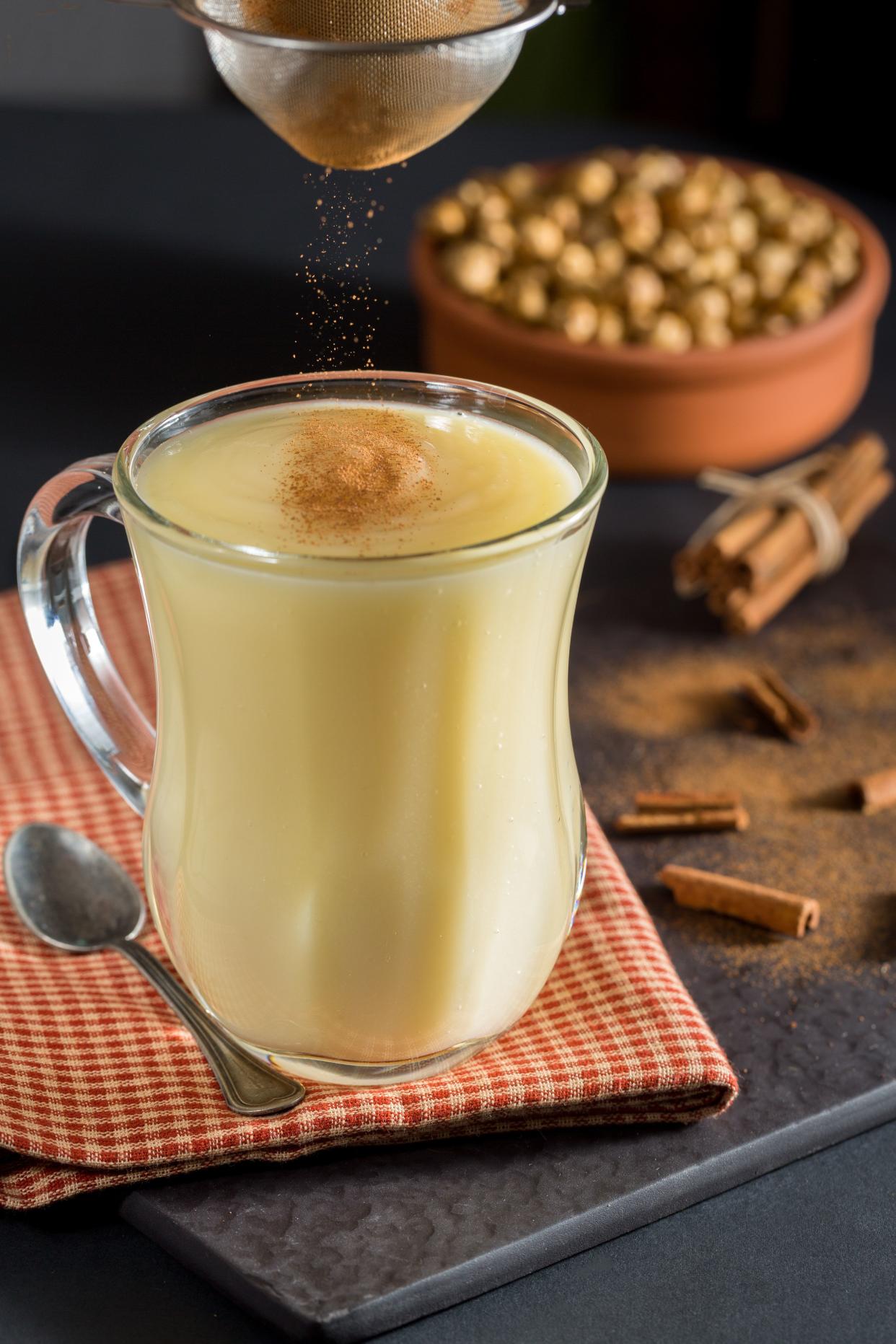 Red and white checkered napkin with a silver spoon and a cup of eggnog with cinnamon being sprinkled into it.