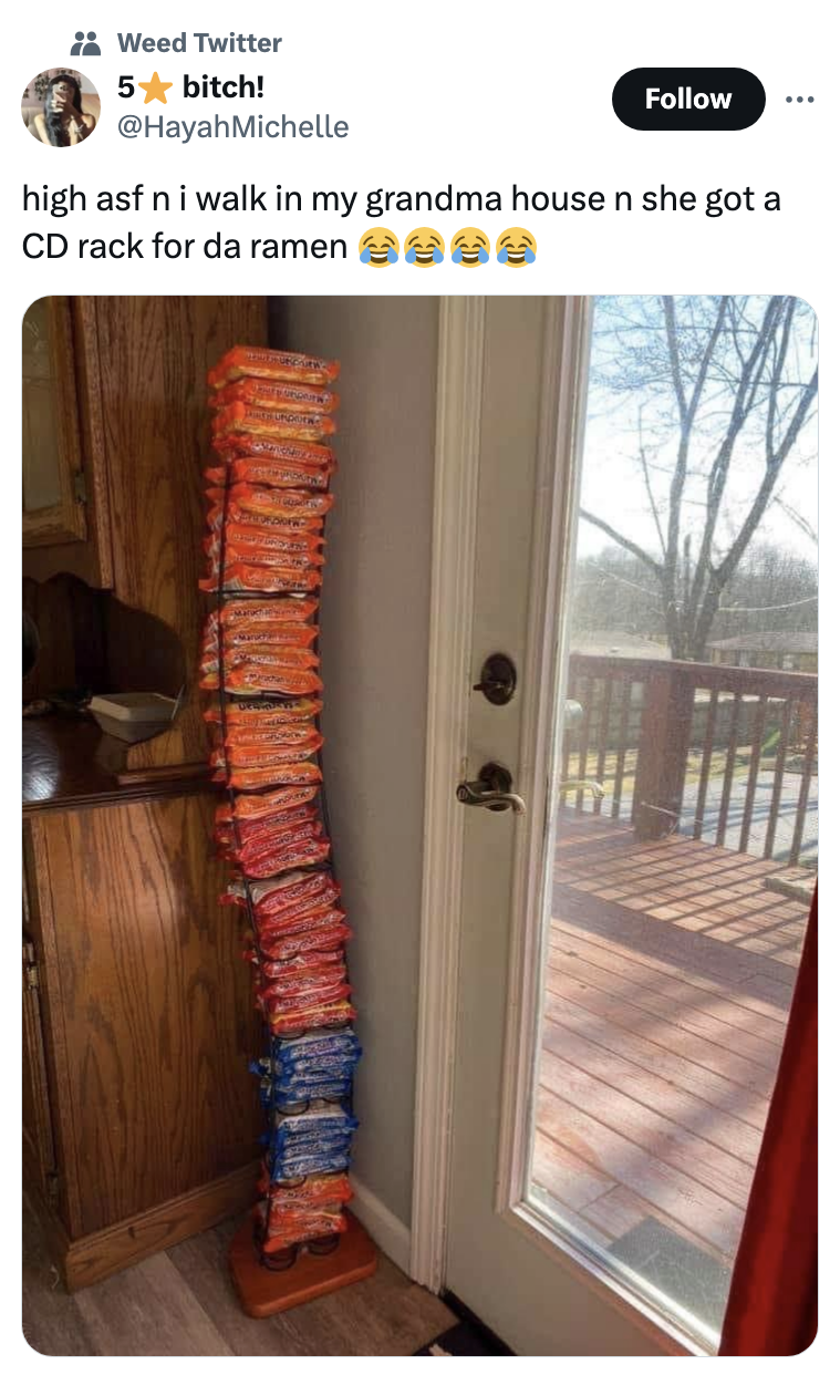 A tall stack of ramen noodle packages is organized on a CD rack next to a door leading to a porch. Tweet by @HayahMichelle humorously notes this organization