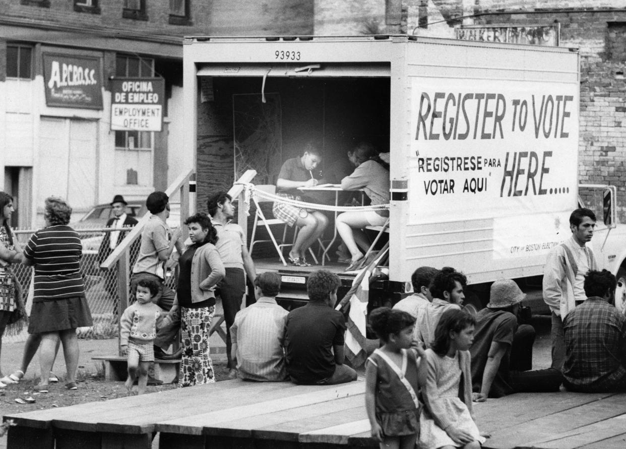 A black-and-white photo shows people gathering around a truck marked with a sign the reads: Register to Vote Here.