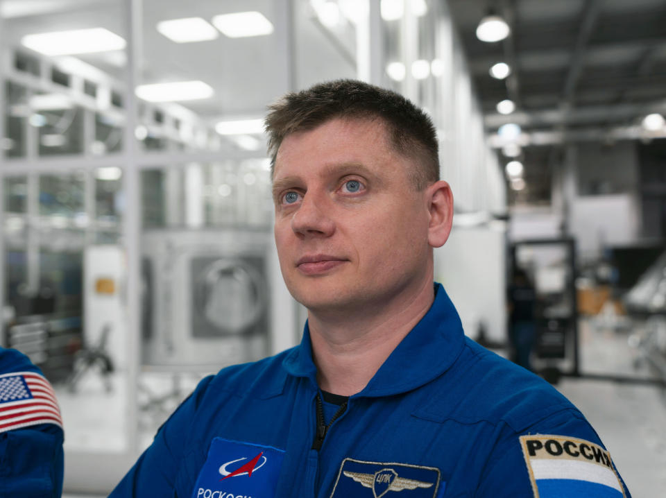 a man in a blue jumpsuit stares into the distance.  He sits in a white room with many glass panels.