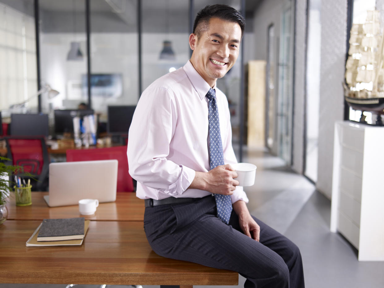 an asian businessman holding cup of coffee sitting on desk in office, smiling and cheerful.