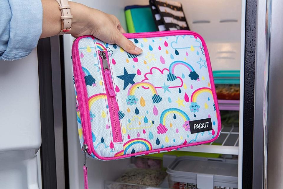 A model's hand placing a bright pink trimmed lunch bag with rainbow designs into a fridge