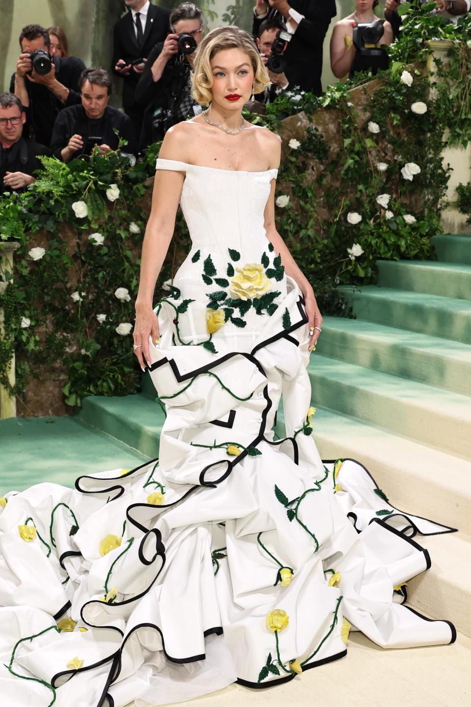 Gigi Hadid in a voluminous white tiered gown with black and green accents, standing on stairs at an event