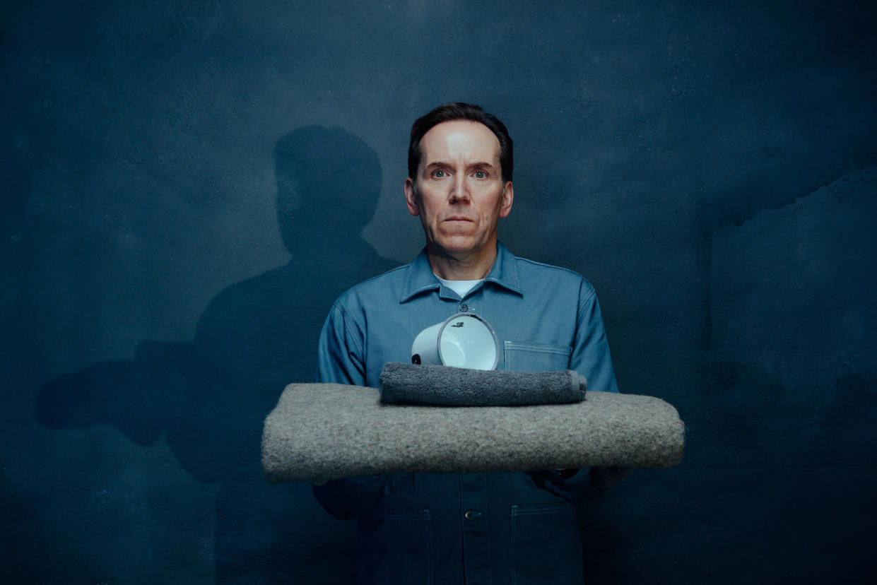  A key art shot from Professor T season 3 showing Professor Jasper Tempest (Ben Miller) in a grey prison overall standing against a charcoal-coloured backdrop. He is holding a folded prison-issue grey woollen blanket in his arms, and on top of that is a small folded black towel and a large white mug. His expression is blank, but slightly startled. 