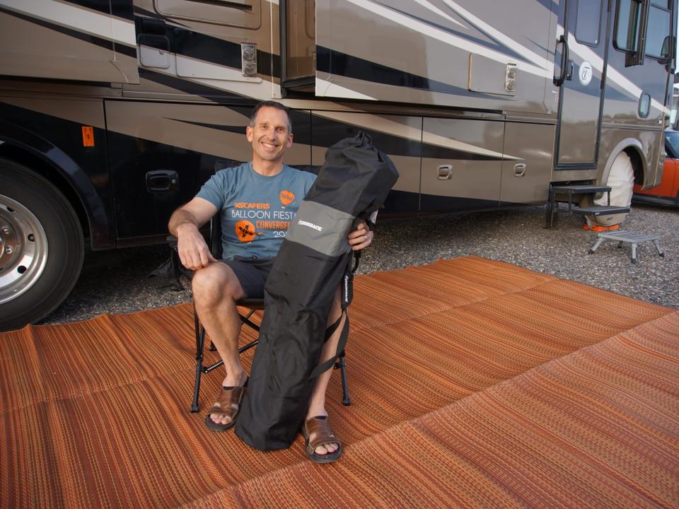 Marc sits on camping chair on a bamboo mat outside the RV with a folded chair in his right hand