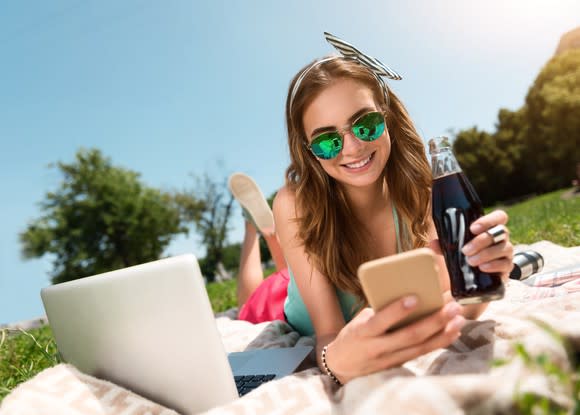 A young woman lying on a blanket outdoors, a soft drink bottle in one hand, a smartphone in the other, and her laptop on the blanket next to her.