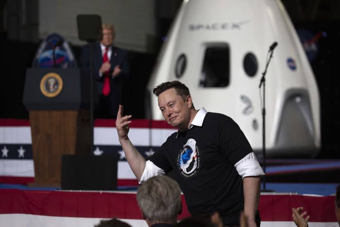 Elon waiving at a Space X event as Donald Trump claps in the background