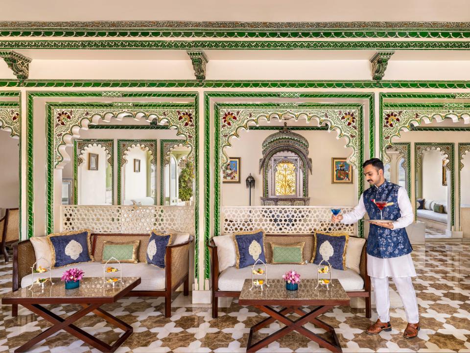 A man carrying colorful drinks in the ornately decorated reception area of the Taj Lake Palace. There are two couches with green, blue, and gold pillows, and a coffee table in front of each one.