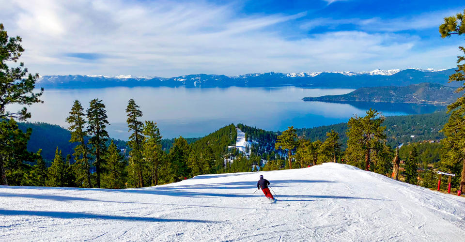 Downhill skiing with Lake Tahoe in the background. A skier in red pants and black jacket is seen from behind heading down a slope towards pine trees.