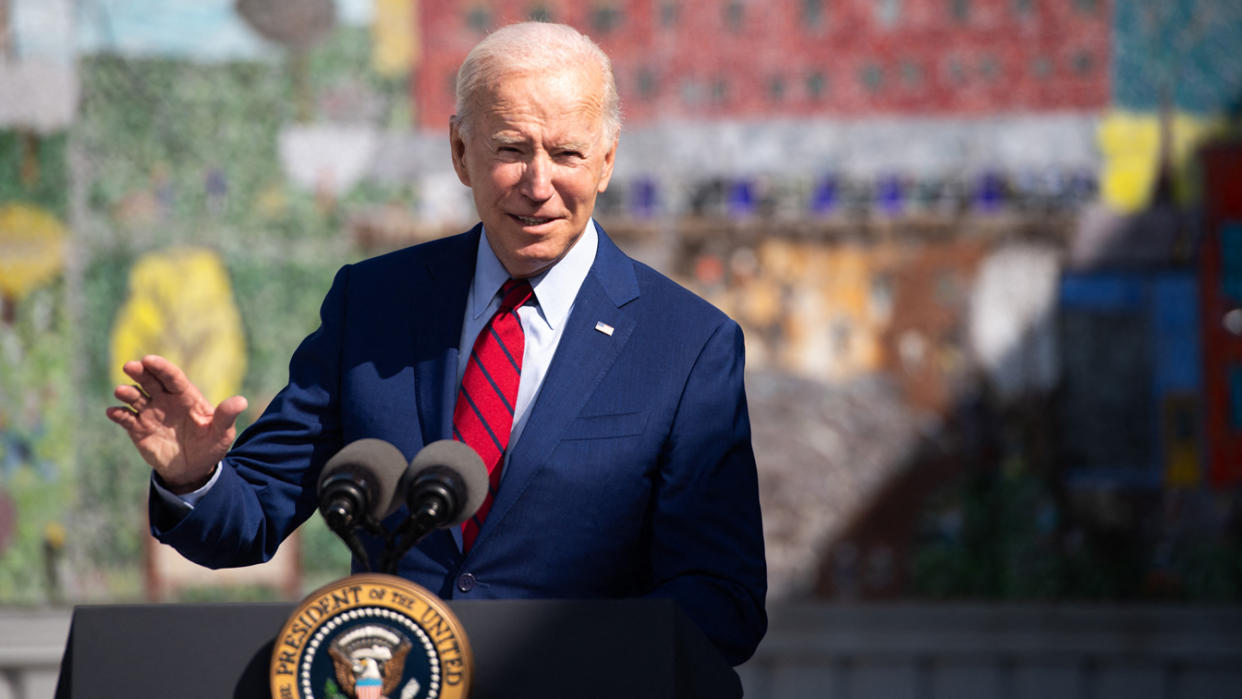 President Biden gestures with his hand while standing at a podium with two microphones and a presidential seal.