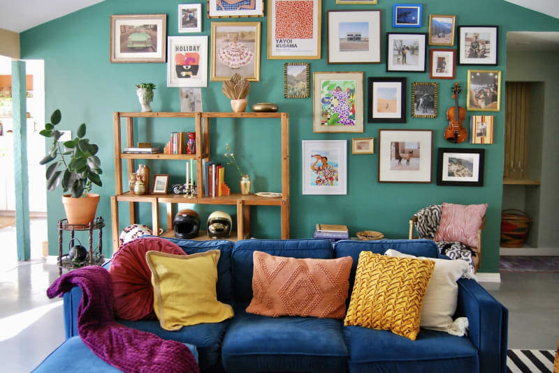 Living room with teal green gallery wall, high ceilings with wood beam, white round pendant light, black and white striped rug, blue velvet sofa with warm colored-accent pillows, cane chairs and coffee table (with glass top), wood shelves with books, art objects