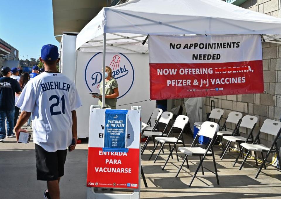 A man in a Los Angeles Dodgers hat and jersey walks past a tent with signs offering free game tickets for each vaccine taken before a game.