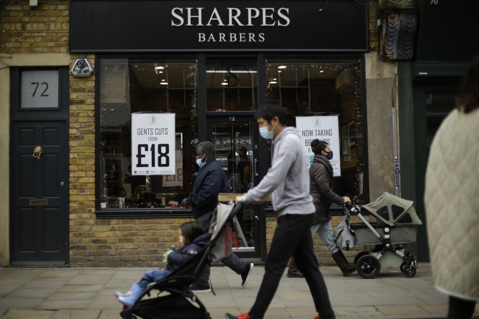 People wearing face masks walk past as barber Matthew Jones stands inside his temporarily closed 'Sharpes Barbers' whilst putting up Christmas lights in the front windows in readiness for when he will be allowed to reopen for business when England's second coronavirus lockdown is due to end on December 2, on Broadway Market in Hackney, east London, Tuesday, Nov. 17, 2020. Small businesses around the world are fighting for survival amid the economic fallout from the coronavirus pandemic. Whether they make it will affect not just local economies but the fabric of communities.(AP Photo/Matt Dunham)