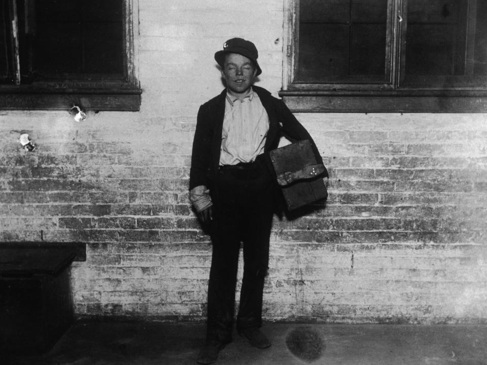 A disheveled shoeshine boy named Tommy, holding a shoeshine kit on a sidewalk in New York City in 1890.