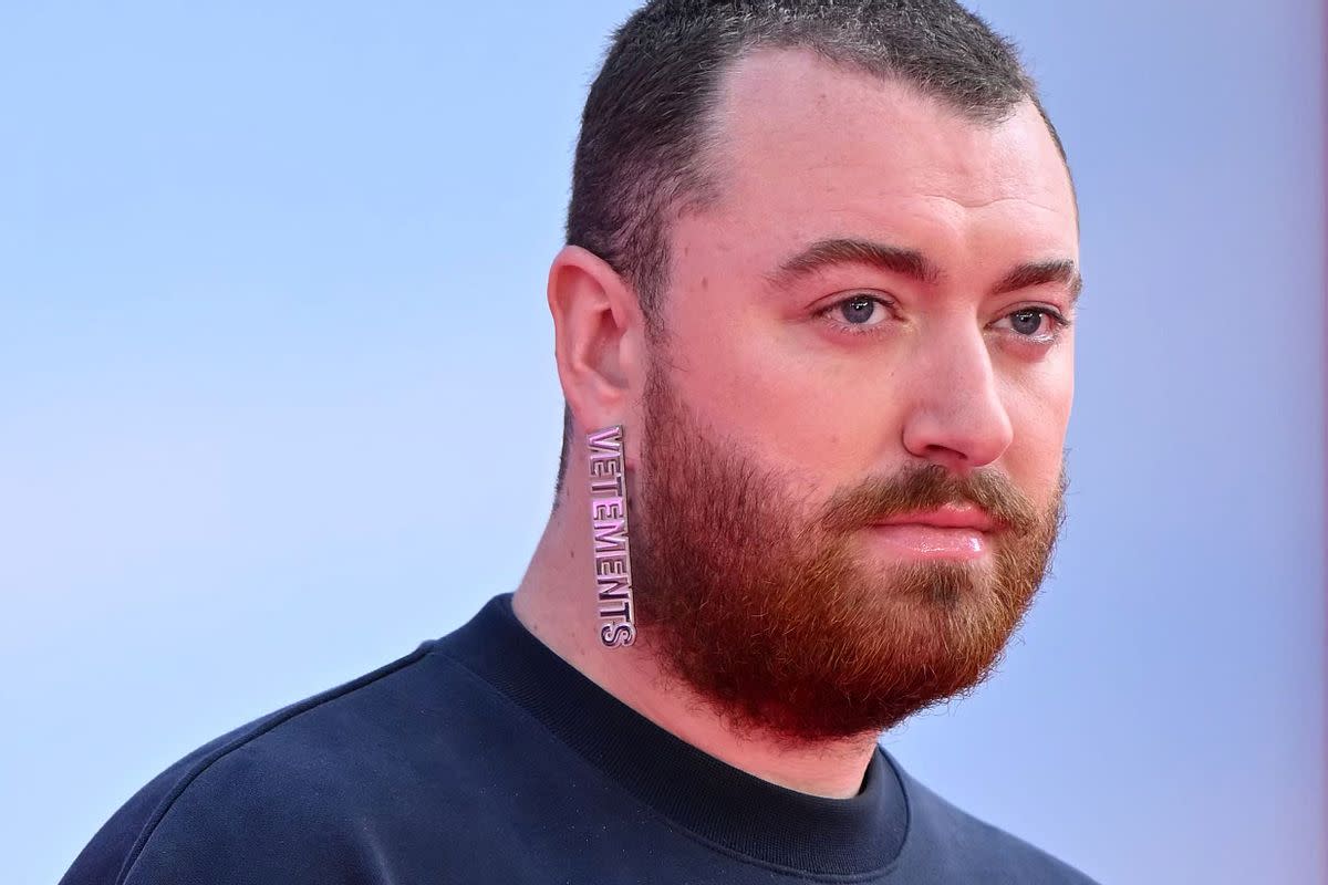 A white person wearing an earring that says "VETEMENTS." They have a beard and are wearing a black top. 