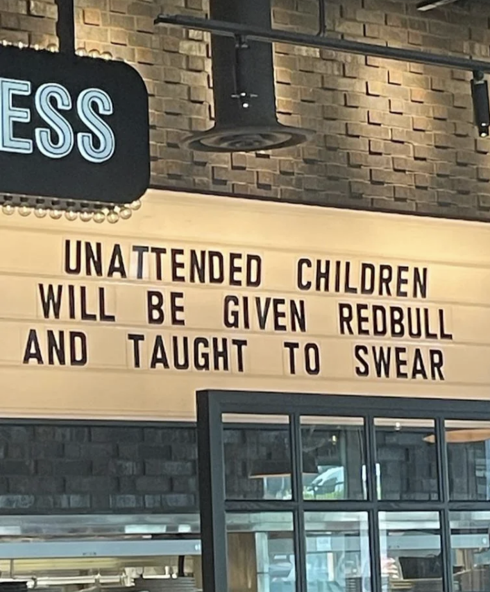 Sign reads: "Unattended children will be given Redbull and taught to swear." Sits above a counter in what looks like a cafe or restaurant