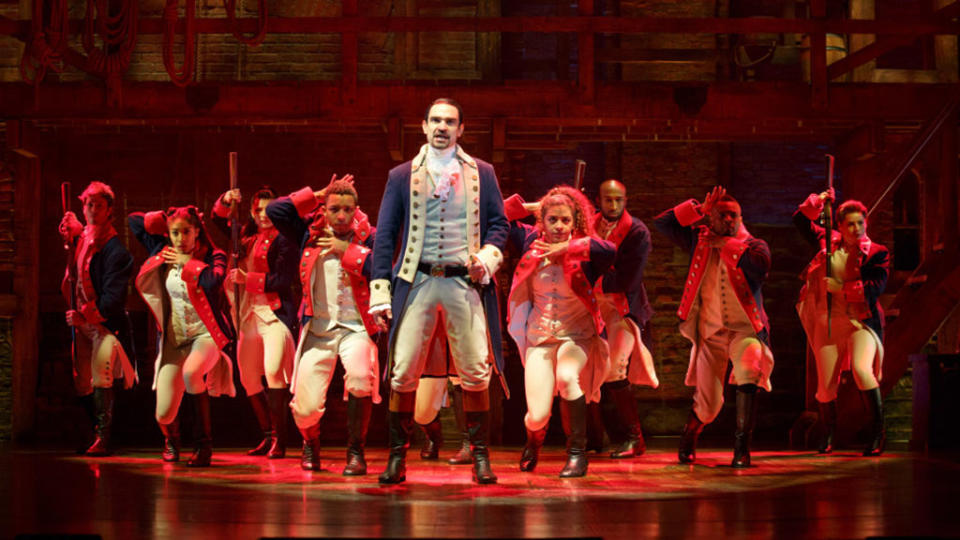 The cast of Hamilton onstage beneath red lighting.