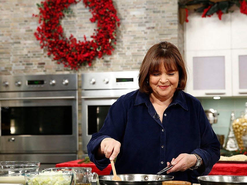 Ina Garten in a kitchen decorated with a red wreath, stirring something in a pan with a wooden spoon.