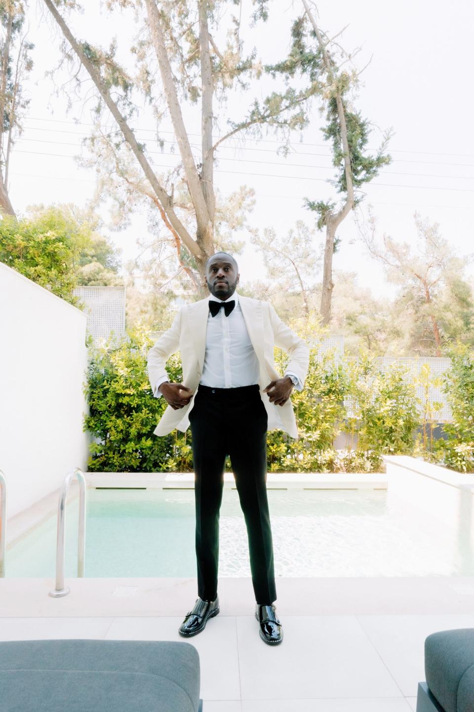 A man in a white tuxedo jacket and black pants stands in front of a pool.