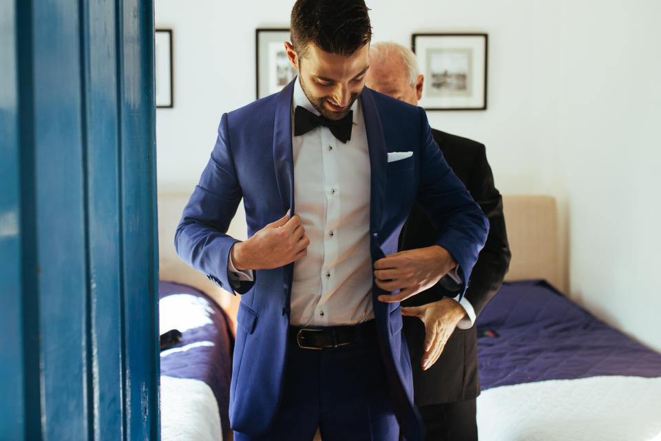 Two men in a bedroom, one adjusting his blue suit jacket while the other stands behind him, helping