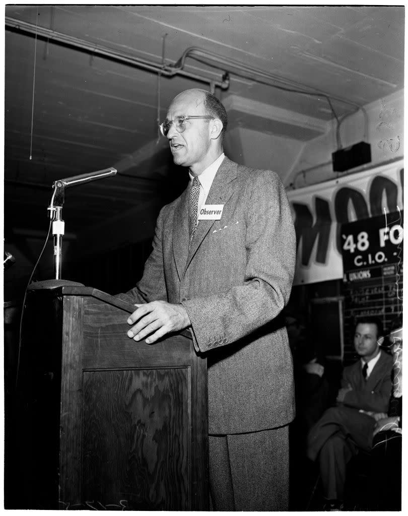 a black and white photo of jimmy roosevelt standing at a podium and speaking into a microphone, wearing a suit and glasses