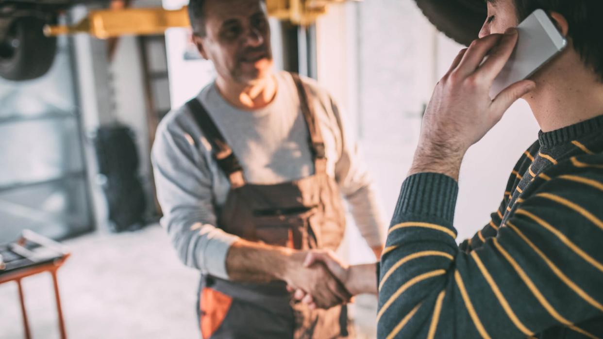 Mature adult auto mechanic, standing under the car, shaking hands with his customer, job is done.