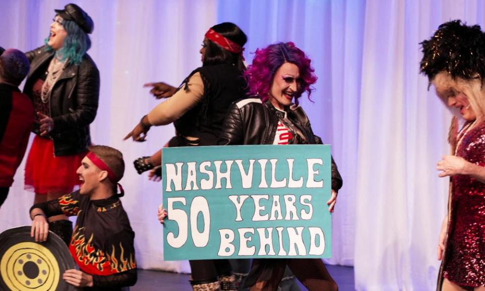 On a stage in front of a white curtain, a handful of drag performers appear to move and speak or sing, one holding a sign that says ‘Nashville, 50 years behind’.