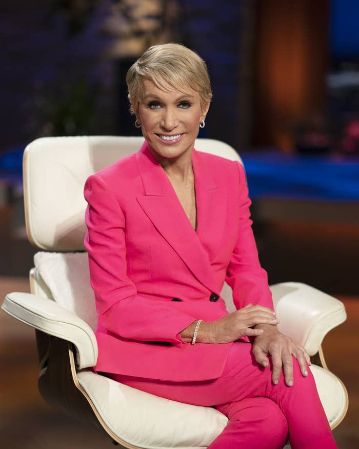Barbara smiles at the camera as she sits on the set of Shark Tank. She's wearing a tailored pantsuit and simple jewelry