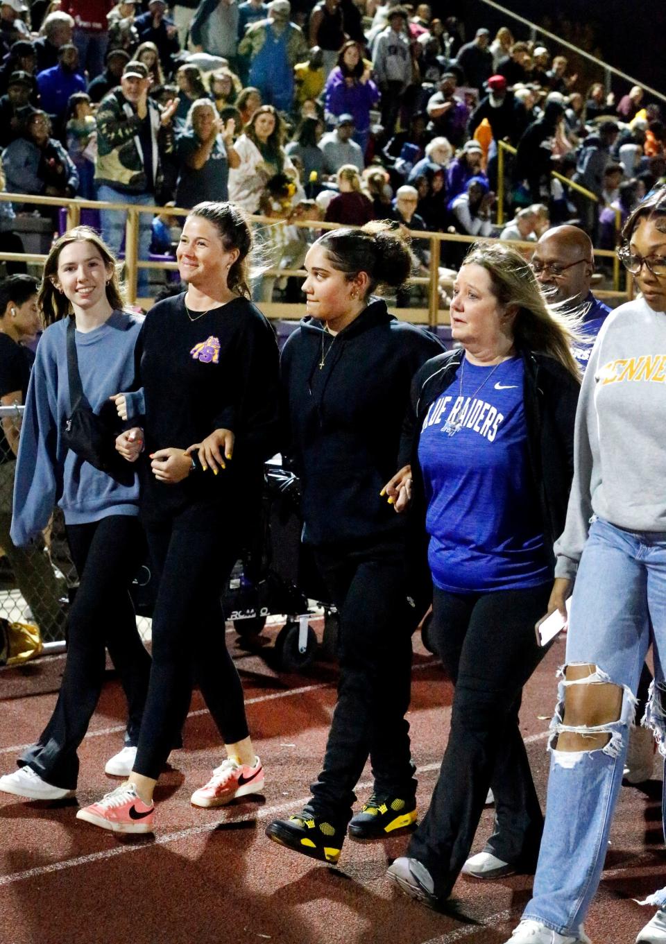 Janae Edmondson, center, walks for the first time in public after losing her legs in an accident less than a year ago along the Smyrna High School sidelines during halftime of the Smyrna football game against Stewarts Creek in Smyrna, Tenn. on Friday, Oct. 13, 2023. Janae Edmondson walks arm in arm with her former high school volleyball coach Katy Bell , left and her mom Francine Edmondson, right. The trio is flanked by former volleyball teammates left Libby Outlaw and Madison Robinson, right. The crowd reacts in the background.