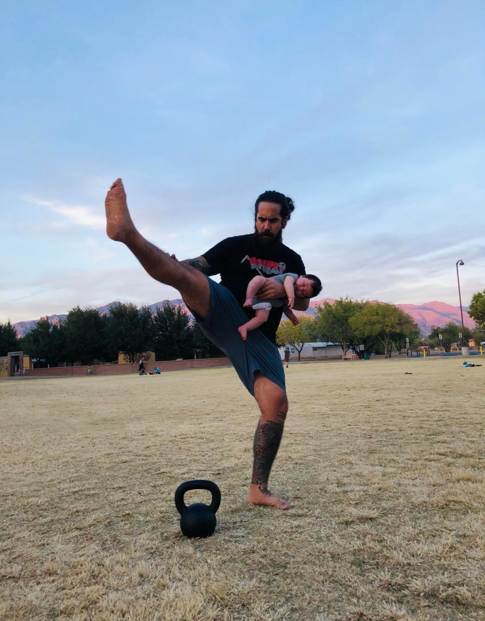 A man with a ponytail holds a baby and exercises outside, kicking up his right leg.