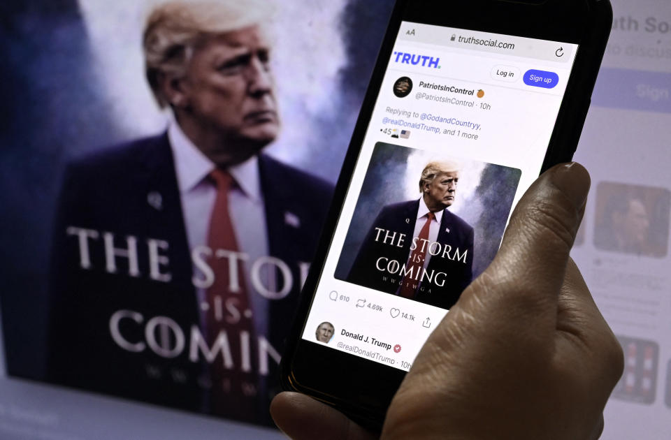 A hand holds a phone displaying a Truth Social post with an image of Donald J. Trump with the message "The Storm Is Coming." Trump is also shown on a computer screen in the background