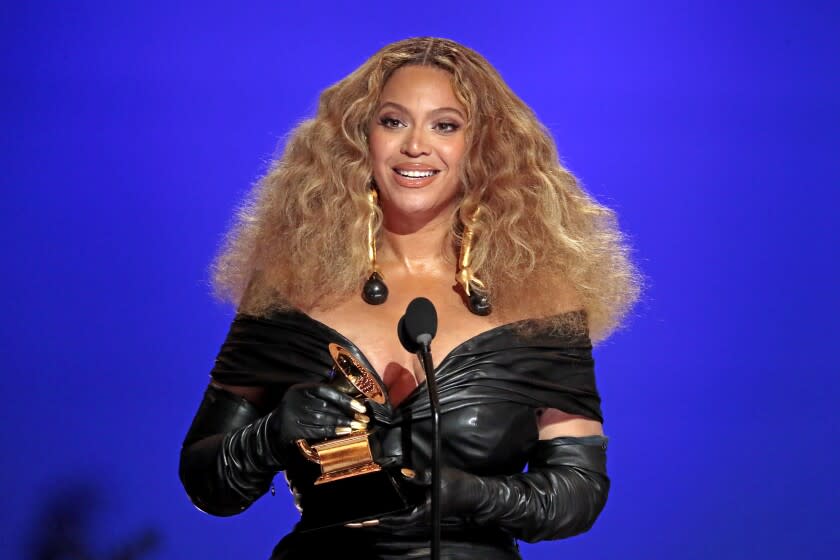 Beyoncé in a black, off-shoulder dress, holding a Grammys trophy in front of a microphone