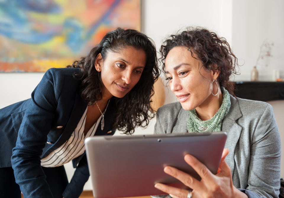 Two businesswomen, one with curly hair and a scarf, the other with wavy hair in a blazer, look at a tablet screen together in an office setting