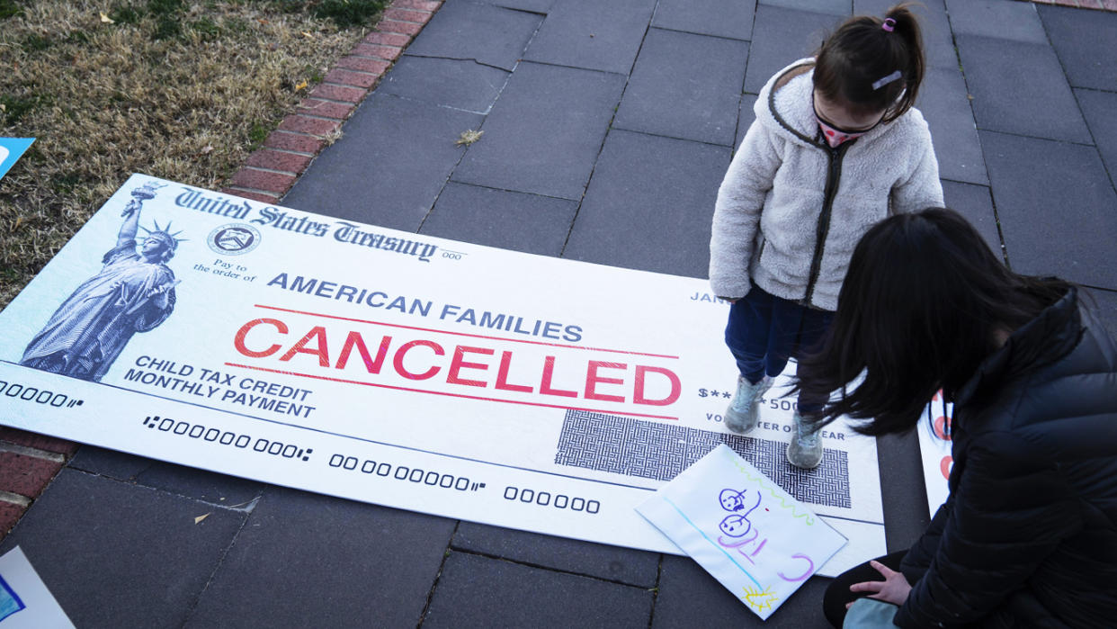 A mother and child on a sidewalk covered with protest signs, including a large one representing a government check made out to American families with the word cancelled emblazoned across it.