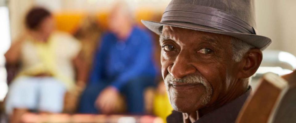 Portrait of elderly black man looking at camera in retirement home, with group of friends in background. Patients relaxing in hospice for seniors.