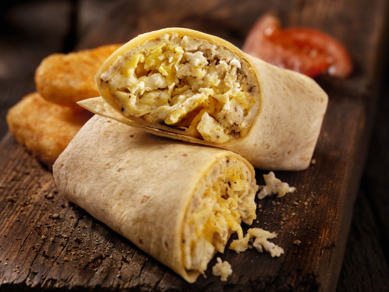 Focus on two halves of a breakfast burrito stacked on each other on a rustic piece of wood with a blurred background of half browns and tomatoes