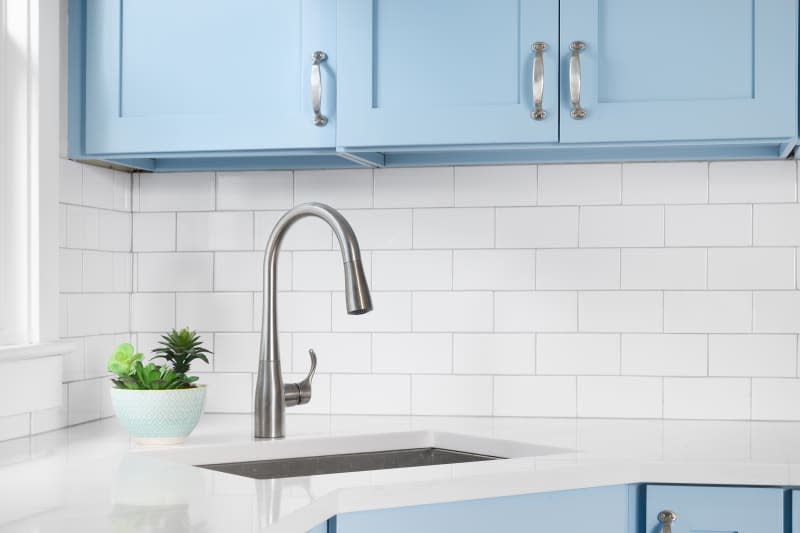 Detail of a kitchen with light blue cabinets, white granite countertop, subway tile backsplash, and a light hanging above a window.