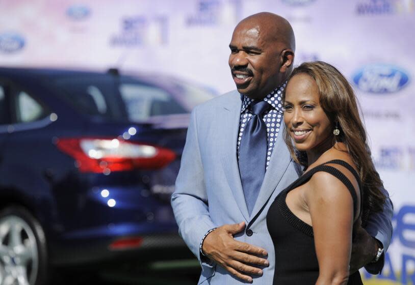 Steve Harvey and Marjorie Harvey smiling and posing together in formal attire in front of a car