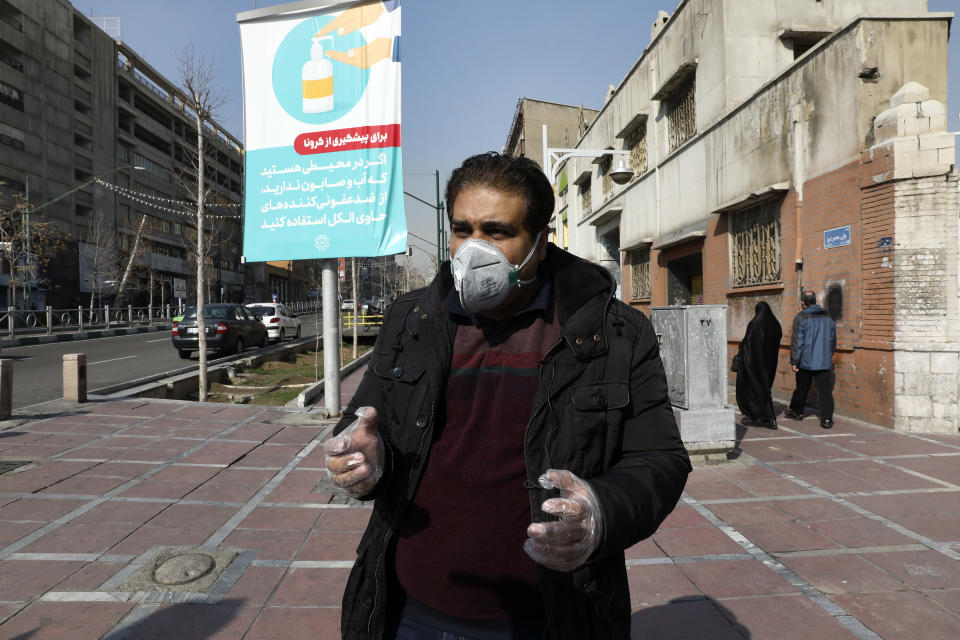 Tehran resident Mohammad Reza Khani, wearing mask and gloves, speaks with The Associated Press in downtown Tehran, Iran, Thursday, Feb. 27, 2020. Amid fear and uncertainty caused by the spread of a new virus, Iranians are taking extra caution to avoid getting infected, as authorities canceled Friday prayers in Tehran, Qom and other cities. The billboard at rear show soap dispenser reading (Farsi) "If you are somewhere with no access to water and soap, use alcohol-based sanitizers." (AP Photo/Vahid Salemi)