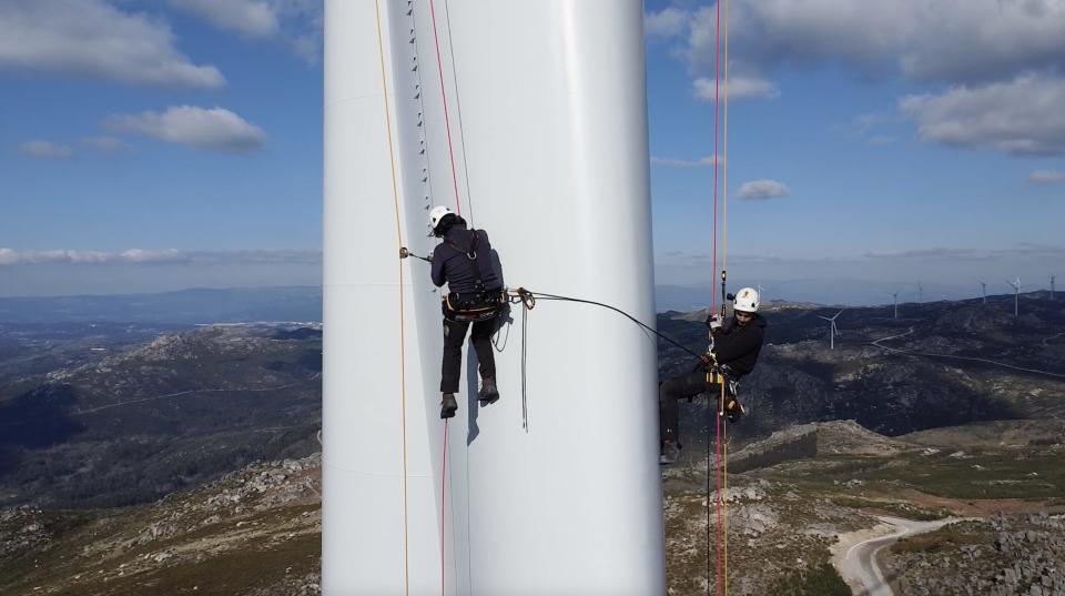 Wind turbine technicians hang from cables with harnesses while inspecting the blade of a turbine.