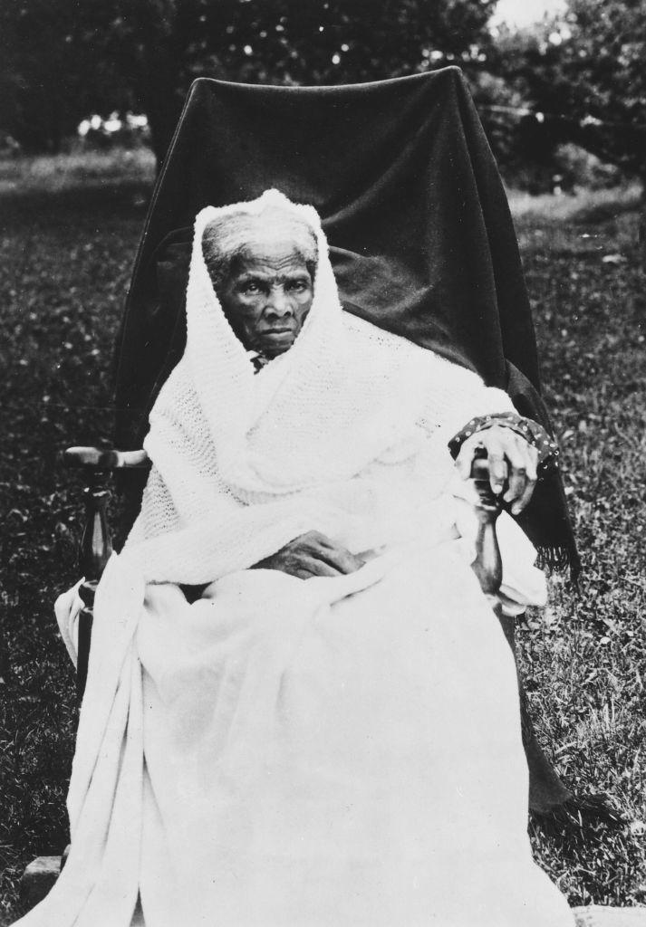 Elderly person seated outdoors, wrapped in a blanket with a solemn expression