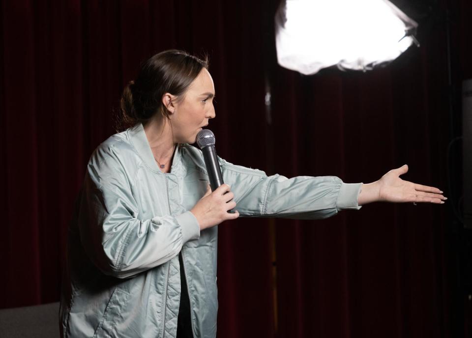 A woman onstage holds a microphone to her mouth and gestures with one arm outstretched.