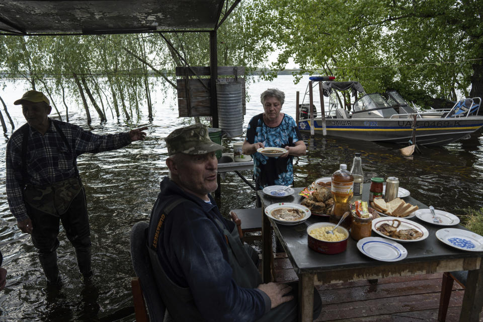 Lyudmila Kulachok, 54, right, sets the food on the table for family diner at the flooded courtyard of her house in the island of Kakhovka reservoir on Dnipro river near Lysohirka, Ukraine, Thursday, May 18, 2023. Damage that has gone unrepaired for months at a Russian-occupied dam is causing dangerously high water levels along a reservoir in southern Ukraine. (AP Photo/Evgeniy Maloletka)