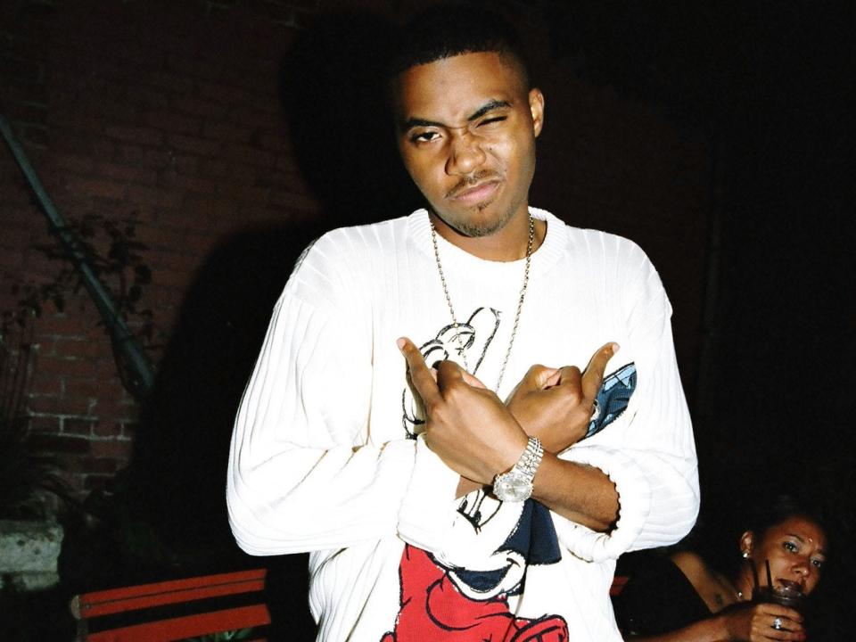 A photo of Nas posing with his middle fingers across his chest while winking at the camera.