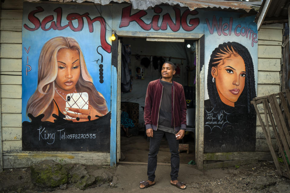 Clovis, 22, poses in front of his hairdressing salon in Goma, Democratic Republic of Congo, Saturday Nov. 26, 2022. At a time of tension and economic uncertainty, the bold names and brightly colored storefronts bring a sense of normalcy to residents who have contended with conflict and natural disasters such as volcanic eruptions for decades. (AP Photo/Jerome Delay)