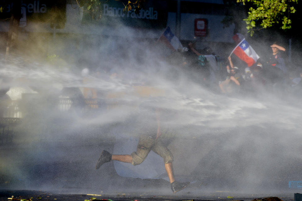 An anti-government runs through spray coming from a police water cannon during clashes in Santiago, Chile, Monday, Oct. 28, 2019. Fresh protests and attacks on businesses erupted in Chile Monday despite President Sebastián Piñera's replacement of eight important Cabinet ministers with more centrist figures, and his attempts to assure the country that he had heard calls for greater equality and improved social services. (AP Photo/Matias Delacroix)