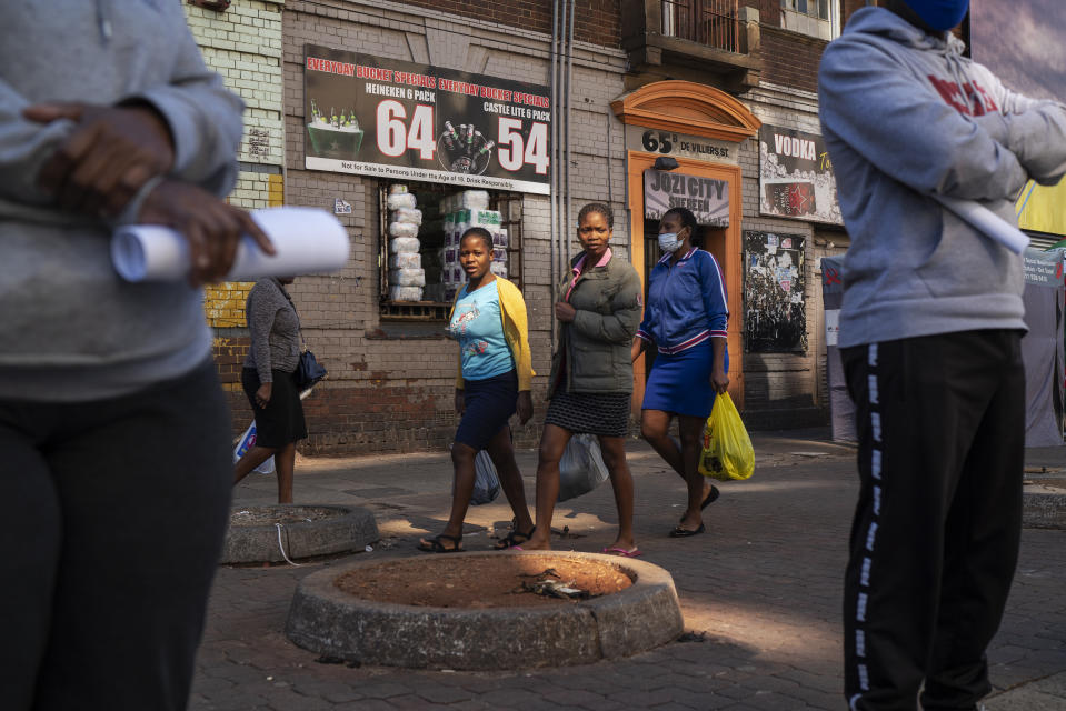 Three women carrying grocery bags walk past residents lining up to be tested for COVID-19 as well as HIV and Tuberculosis, in downtown Johannesburg Thursday, April 30, 2020. Thousands are being tested in an effort to derail the spread of coronavirus. South Africa will began a phased easing of its strict lockdown measures on May 1, although its confirmed cases of coronavirus continue to increase. (AP Photo/Jerome Delay)