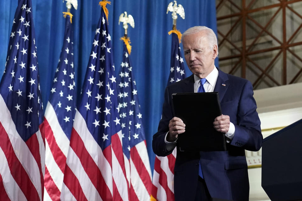 President Biden looks down while holding a black folder and standing near a podium and American flags.